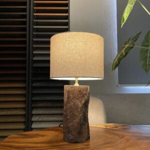 Hình ảnh thực tế đèn bàn gỗ tự nhiên Forest Relic DT-0023 đặt trên bàn gỗ. Sản phẩm có thân trụ màu nâu với bề mặt mộc, đường vân và vết nứt tự nhiên, kết hợp chao vải hình trụ màu kem. Đèn sử dụng bóng E27 và nguồn điện 220V–50Hz.